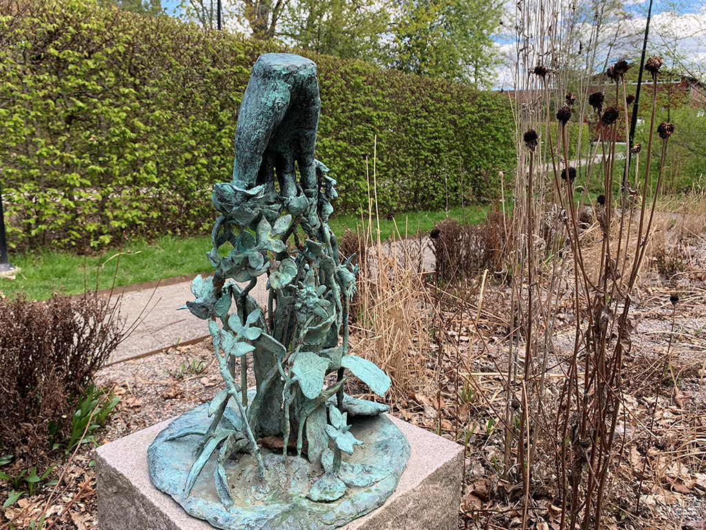 Bronskulptur av en hand som doppar fingrarna i blommor. Skulpturen finns i en park med en grönskande häck i bakgrunden.