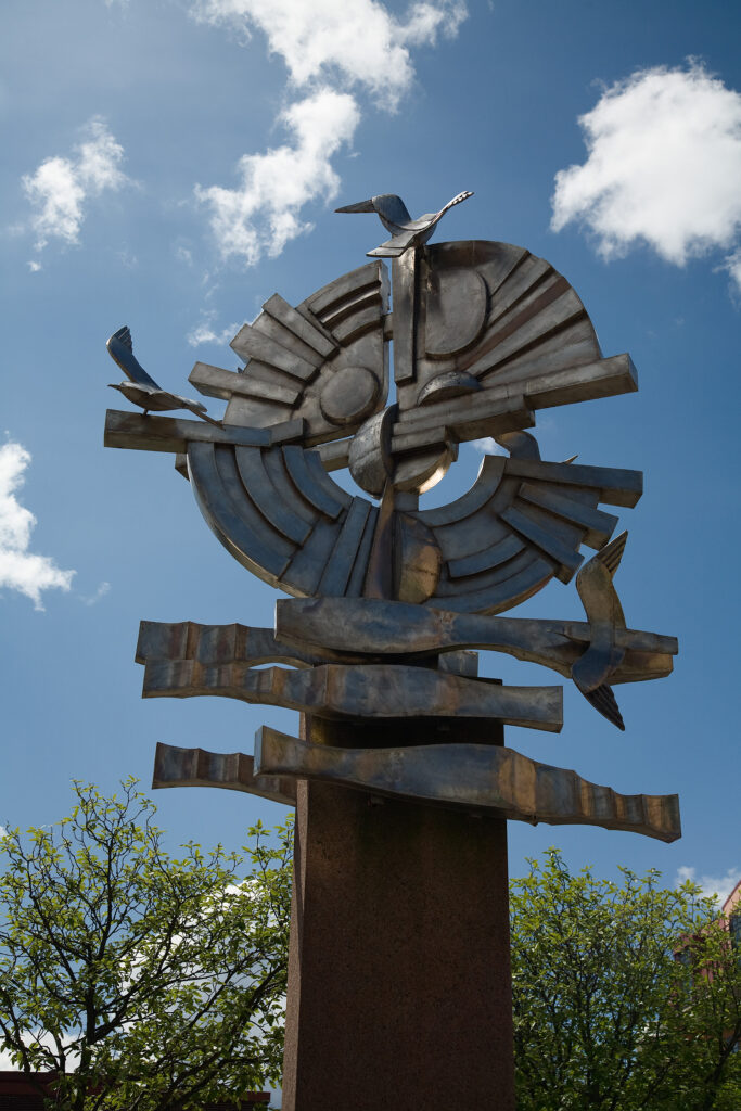 Skulptur av en rund form av stål på en sockel mot en blå himmel. På rundeln sitter fåglar av stål.