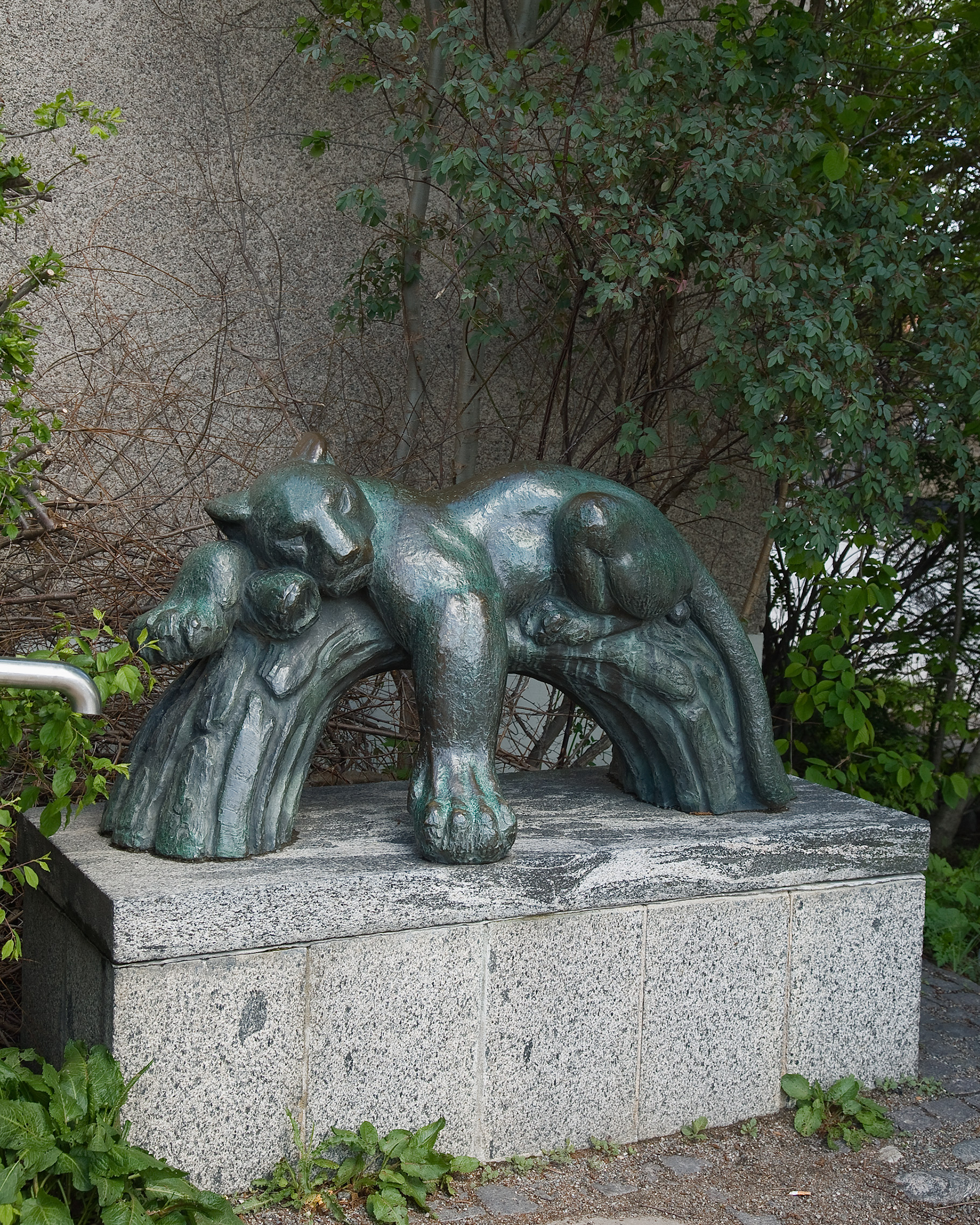 En skulptur av en panter vilar på en gren i brons. Skulpturen står på en grå sockel i ett buskage.