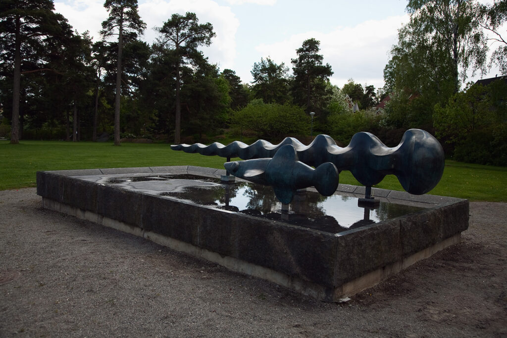 Två skulpturer i en vattenfylld bassäng i en park. Den ena är en abstrakt avlång vågig form, den andra en fisk.