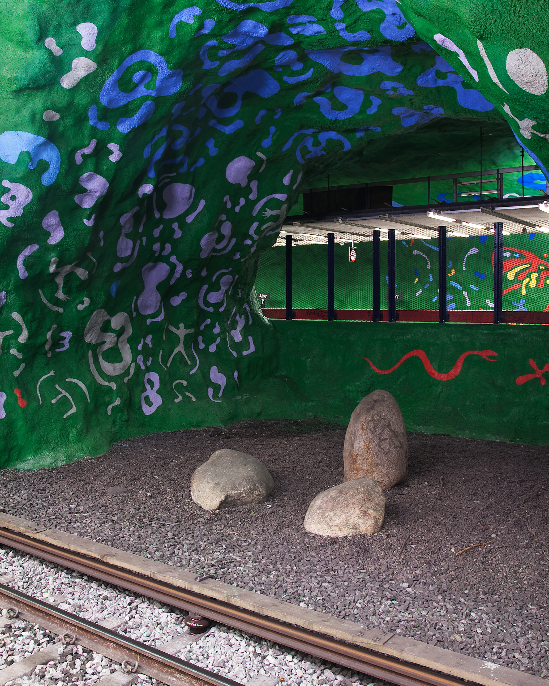 Målade väggar som i en grotta. Blått mönster på grön botten. På marken en räls, grus och stenar.