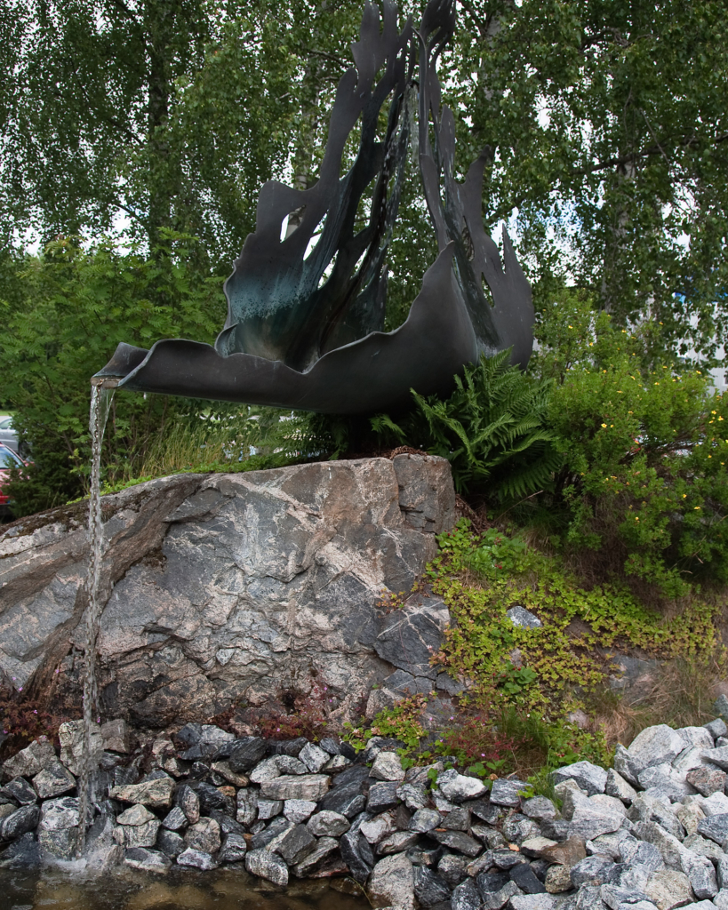 Abstrakt grönaktig skulptur som det rinner vatten ur ner i en damm med stenar. Skulpturen står på en sten med grönskande skog runtom.