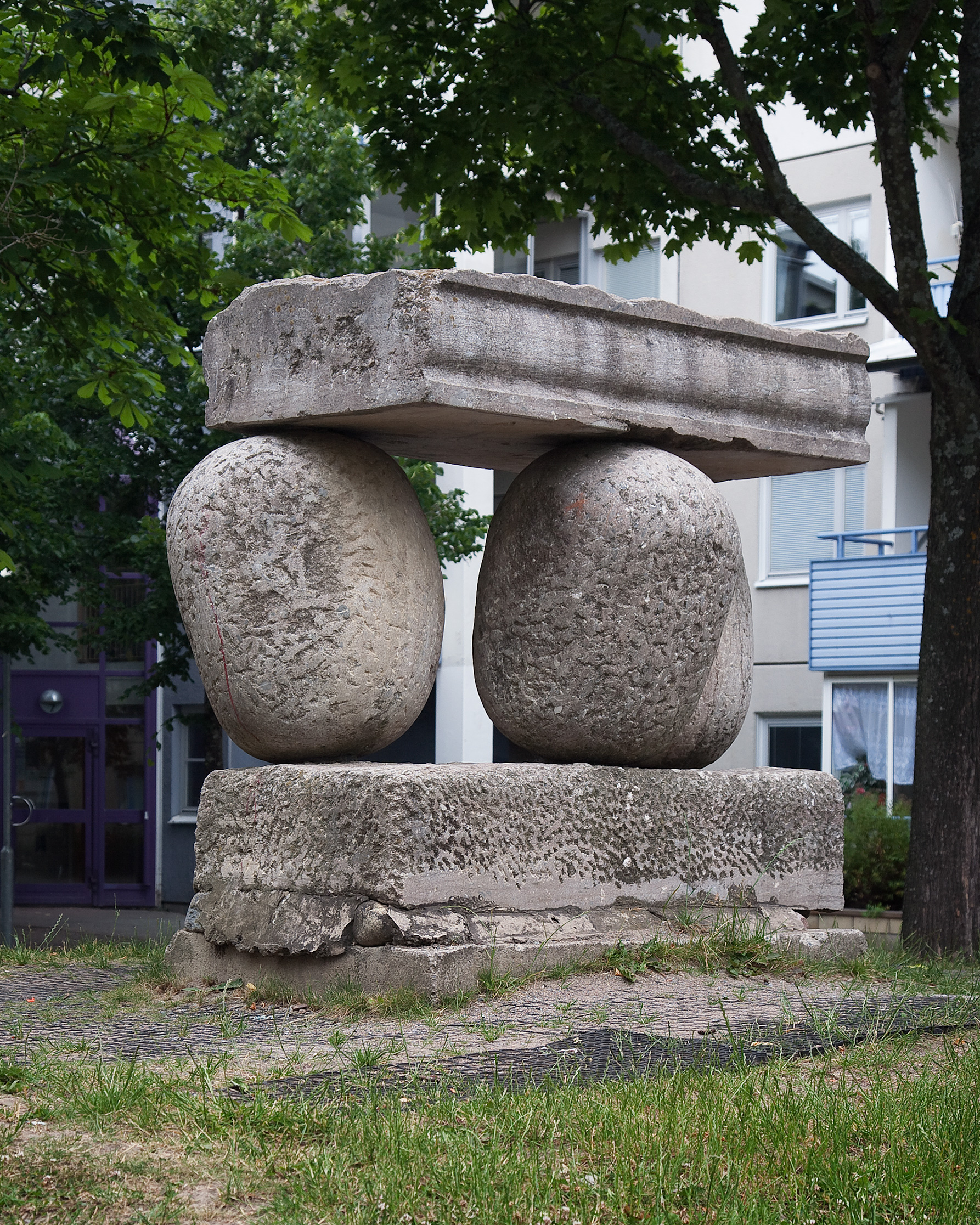 Skulptur med två rundade stenar som balanserar på ett grått stenblock med ett stenblock ovanpå. Gräs framför och träd och hus bakom