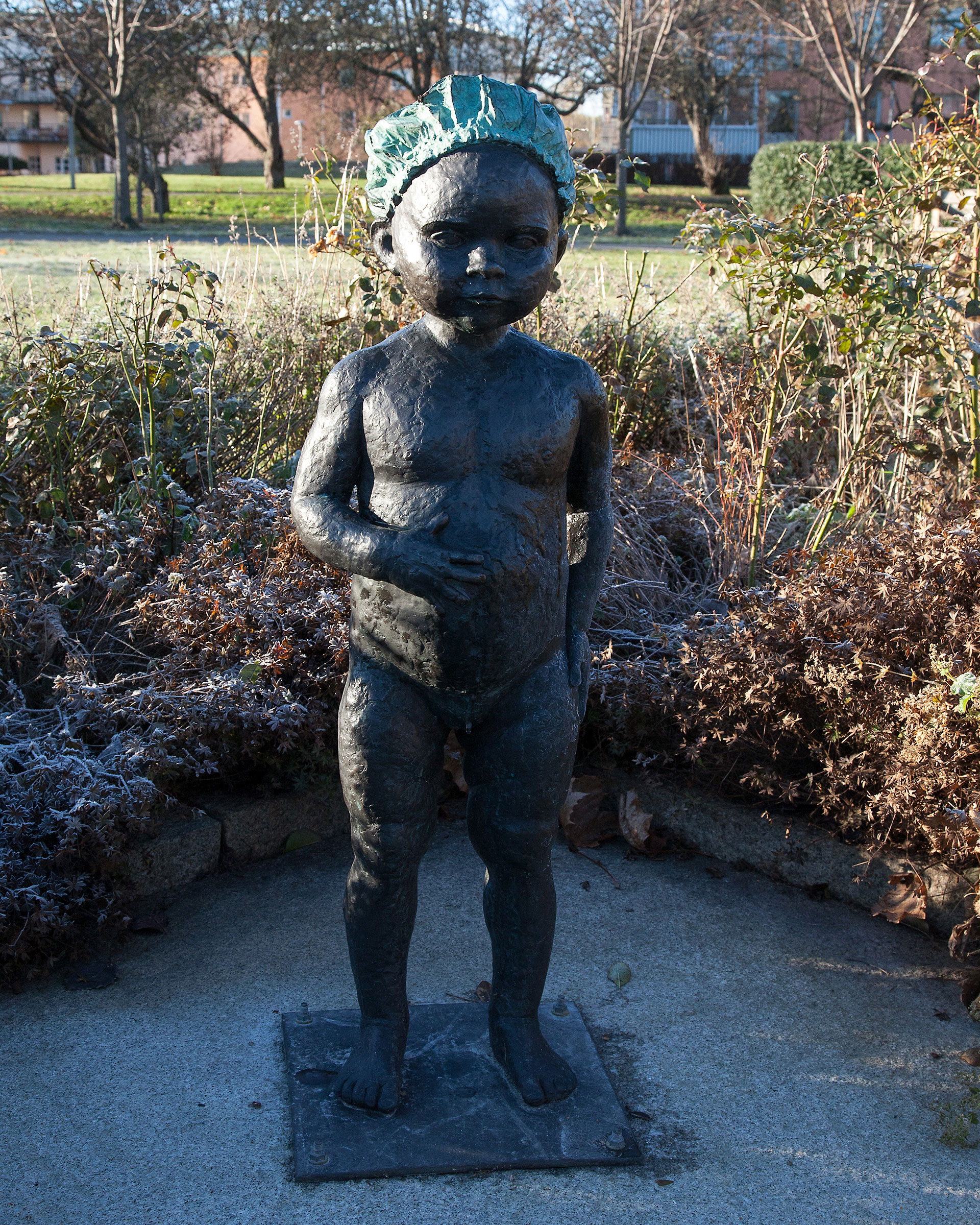 Ett skulptur av ett naket barn som håller handen på magen och  står framför en buske.