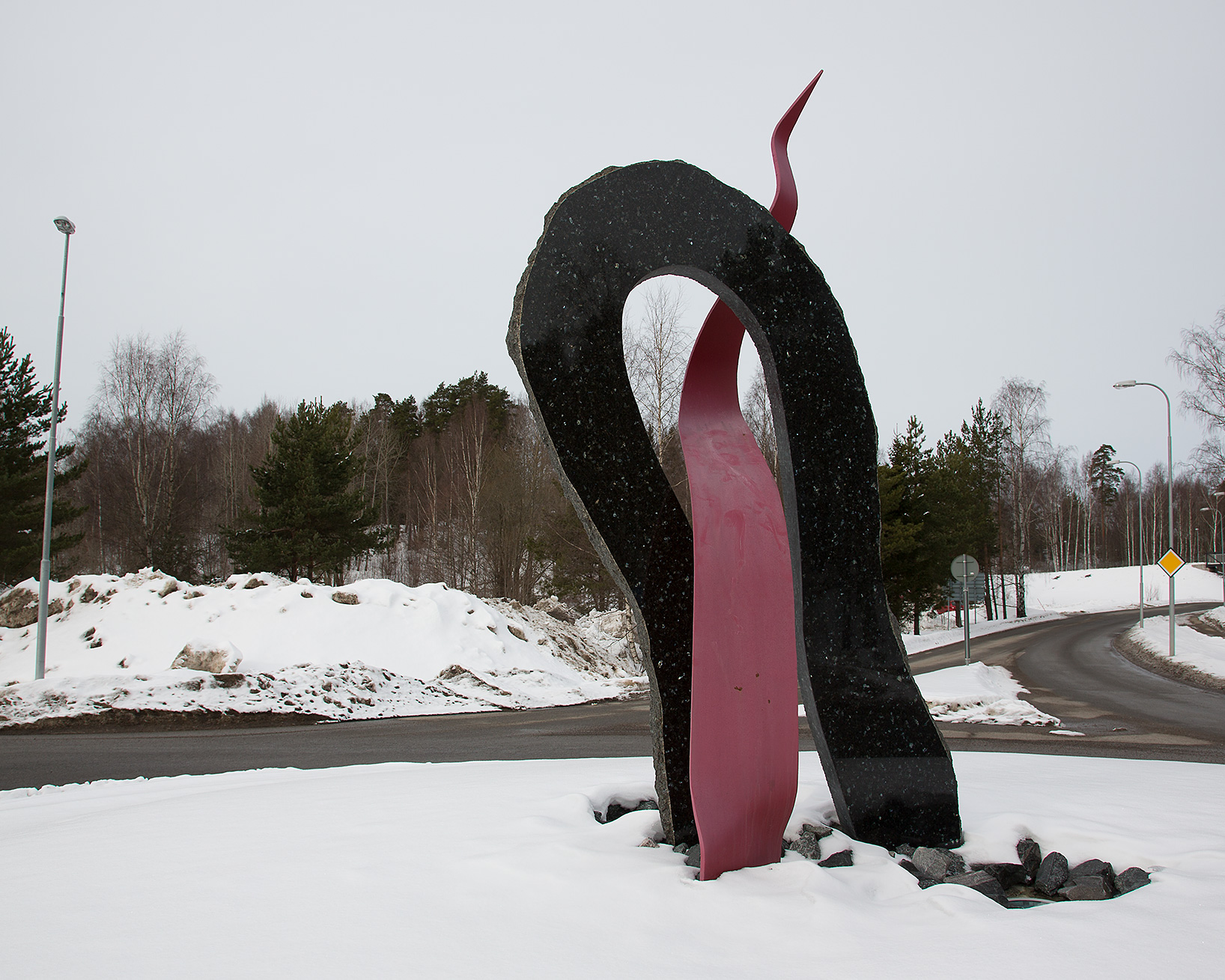 EEn svart hög hålform som står på marken. En röd abstrakt tungliknande form går igenom den svarta formen. Snö och vägar.