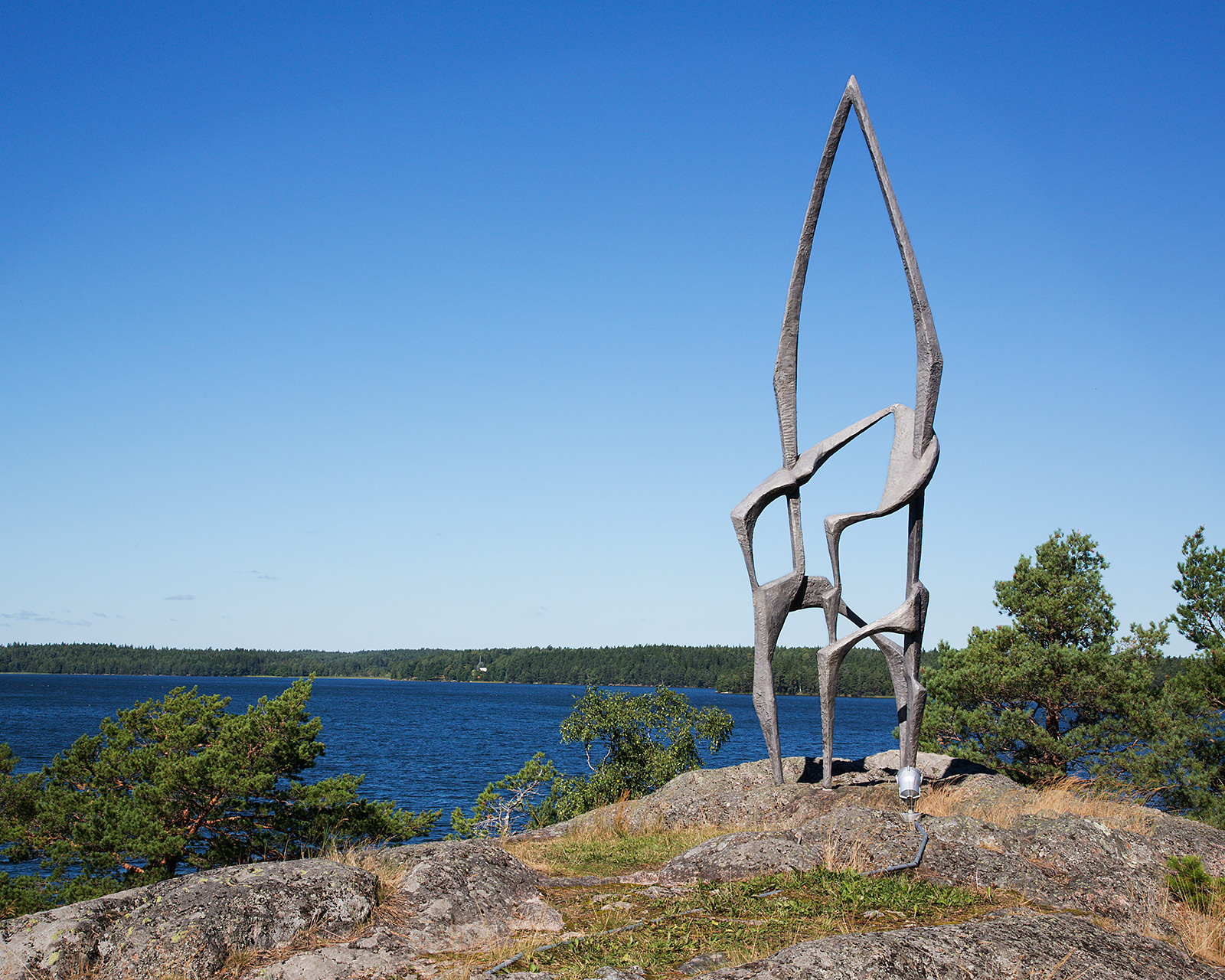 En figur av metall som liknar en stol med en spetsig båge upptill på en klippa vid vatten.