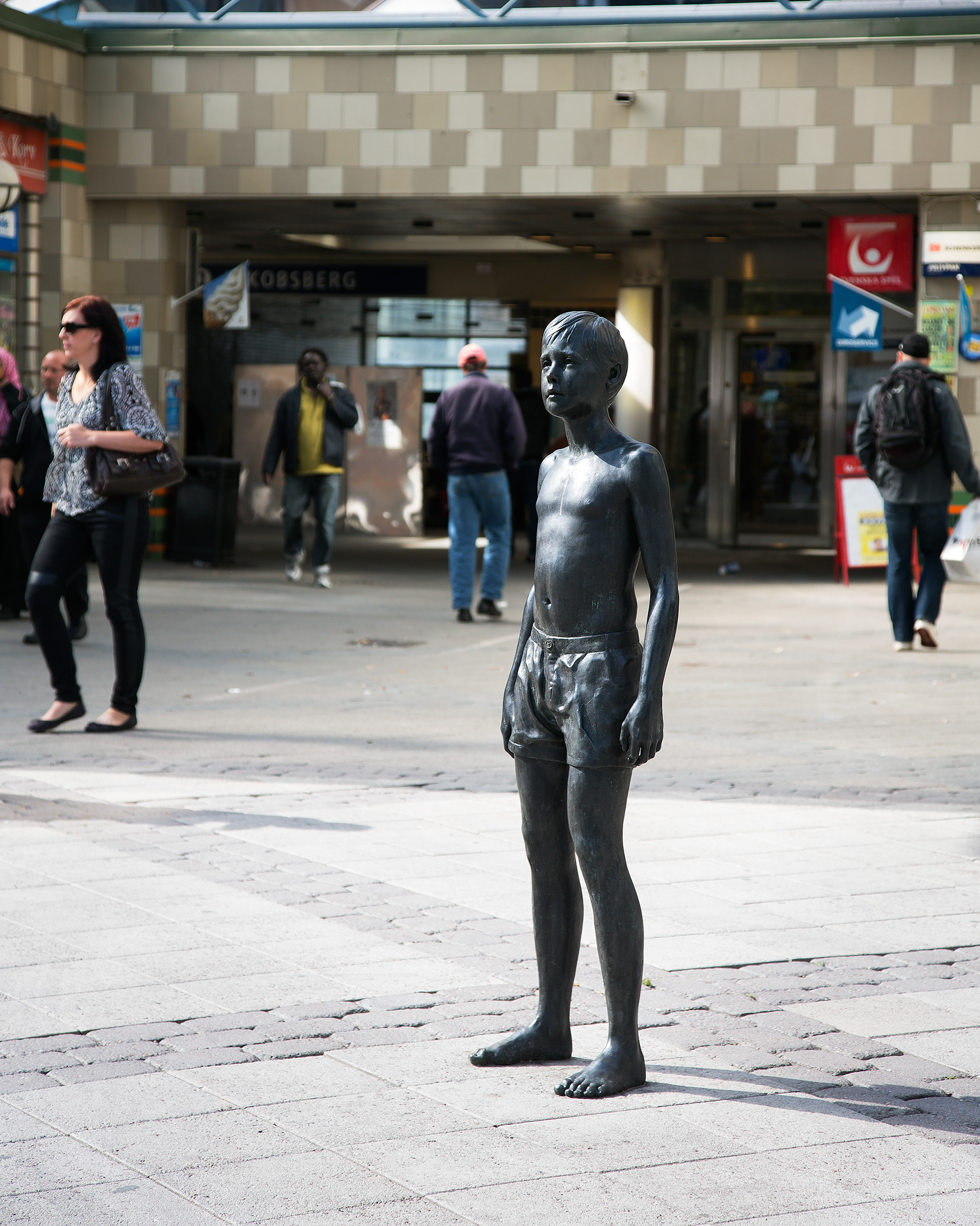 En skulptur i brons av pojke i shorts står i stadsmiljö. Människor och affärer bakom.