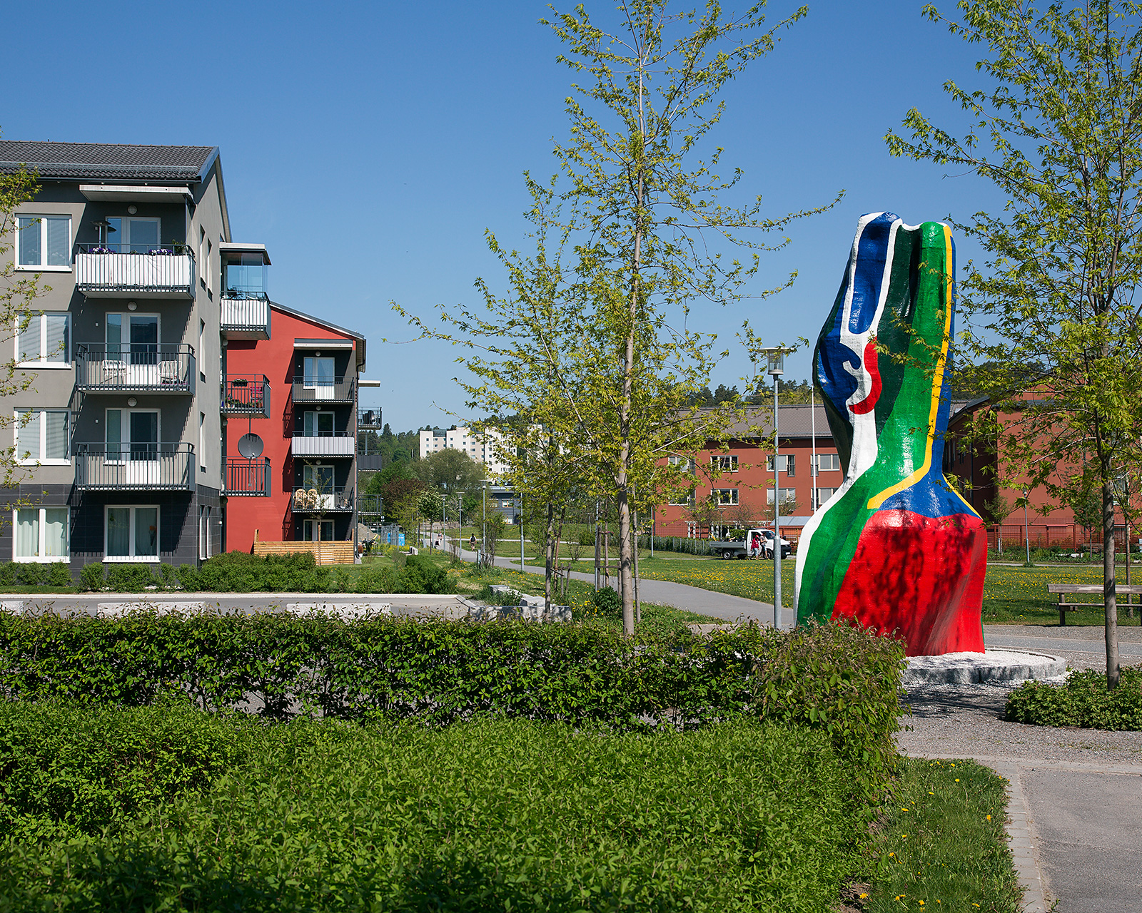 En abstrakt skulptur i färgerna blått, rött, grönt, vitt. Flerbostadshus bakom. Grönskande träd och häckar runtomkring.