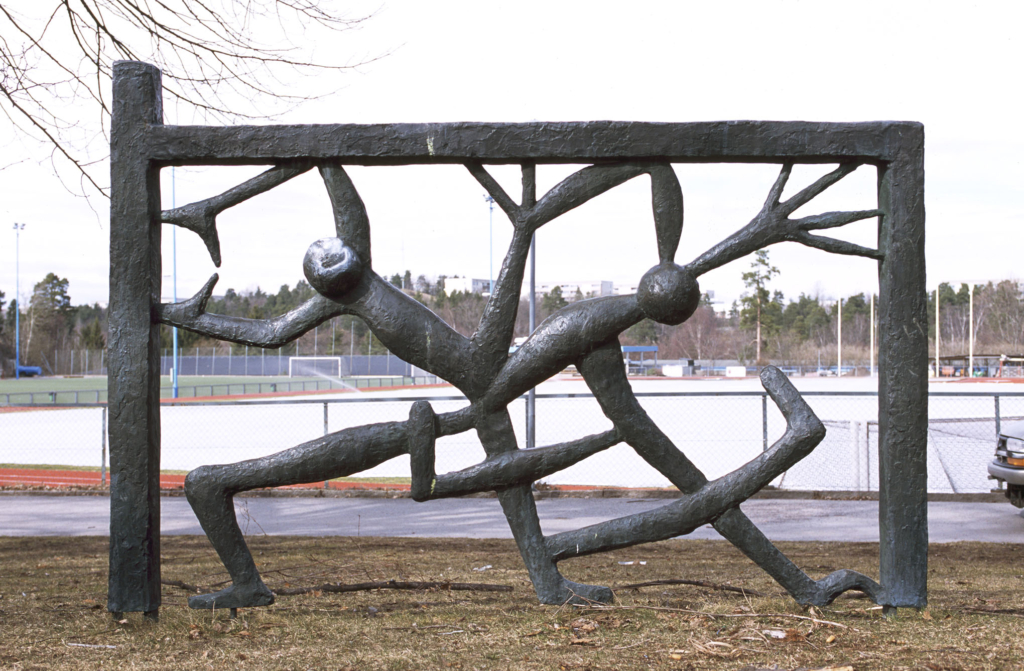 Skulptur i brons med en fyrkant och två figurer med huvuden vars armar ben och ben bildar ett mönster. Bakom syns en idrottsplats med ett fotbollsmål.