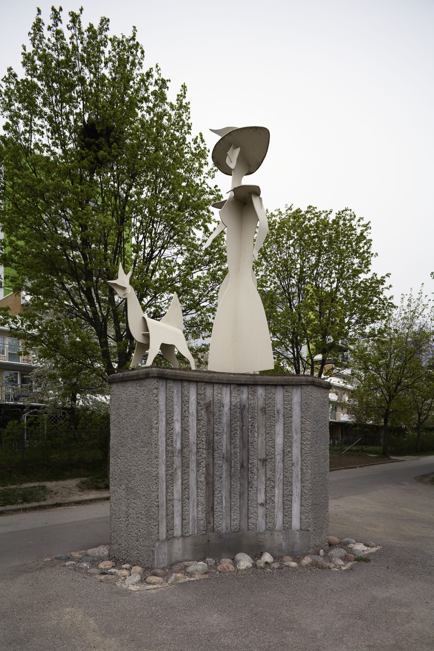 En vit skulptur av en dam står på en hög grå sockel, framför sig har hon en hund.