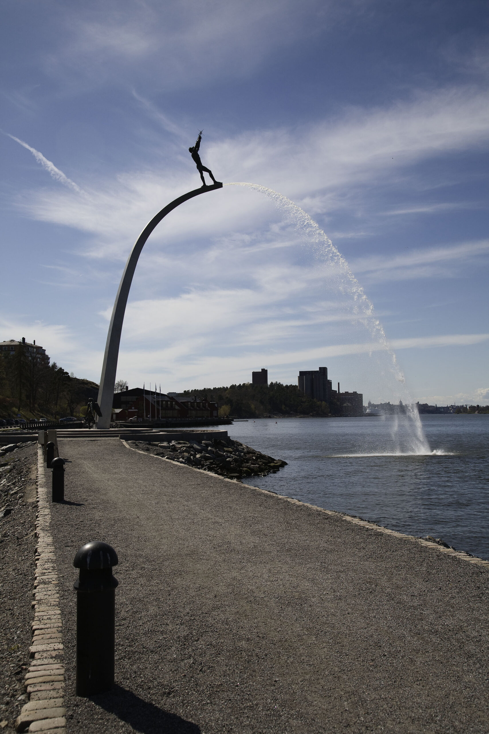En båge över vattnet som sprutar vatten ur sig. Längst upp på bågen står en skulptur av en man. Silhuett av bebyggelse bakom.