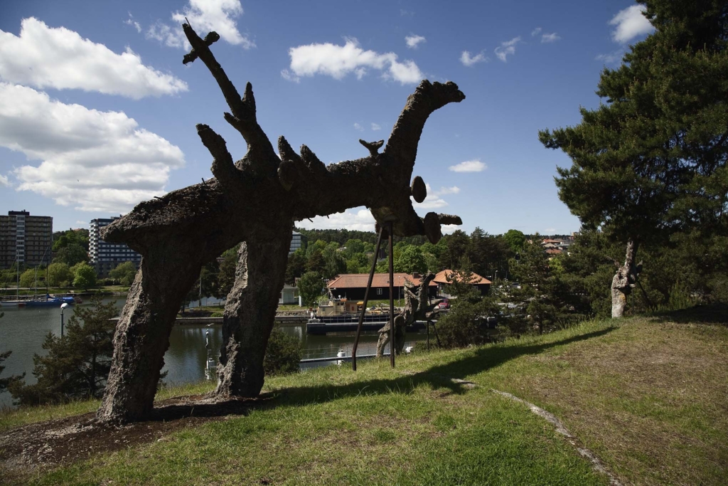 En stor abstrakt skulptur på två ben som står i en grässlänt ner mot vatten. Bebyggelse och träd i bakgrunden.