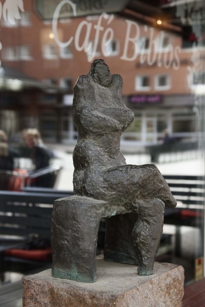 En skulptur av en liten figur som sitter med korslagda ben och armar på en enkel bänk utan ryggstöd.
