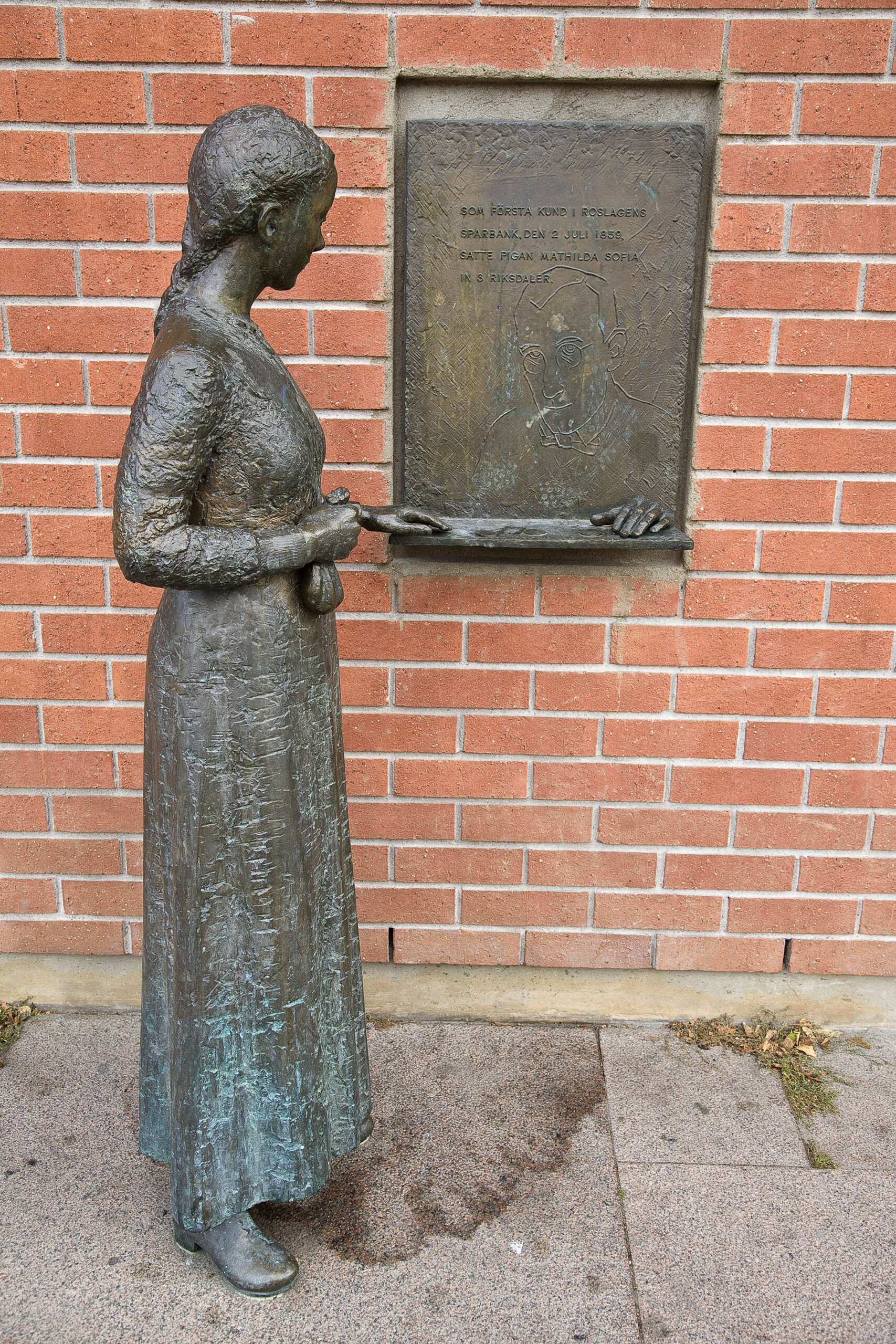 En skulptur av en kvinna i klänning framför en röd tegel vägg och en skylt av brons.