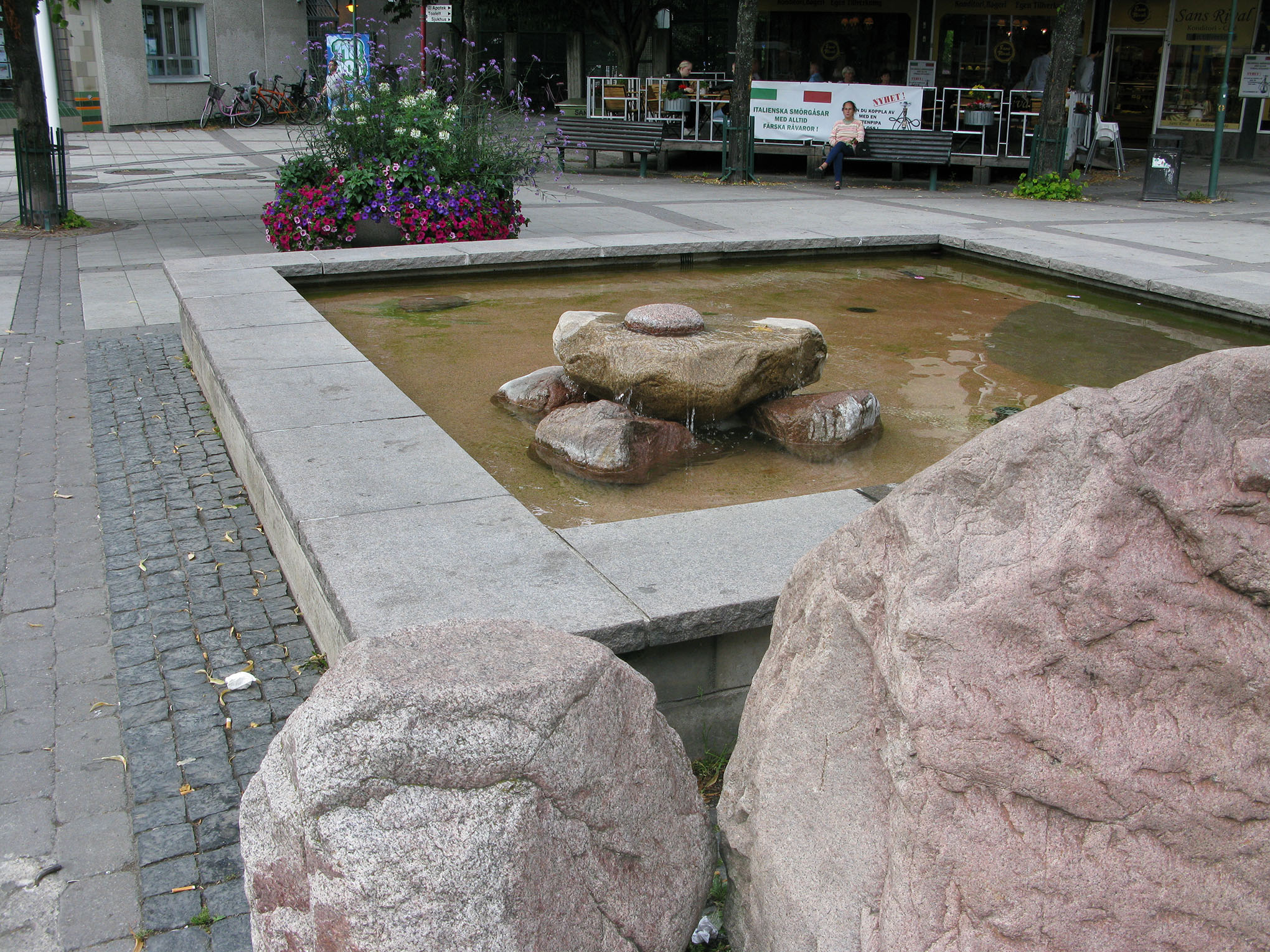 En hög med stenar ligger i en fyrkantig vattenfylld bassäng. Framför bassängen är två stora stenar.