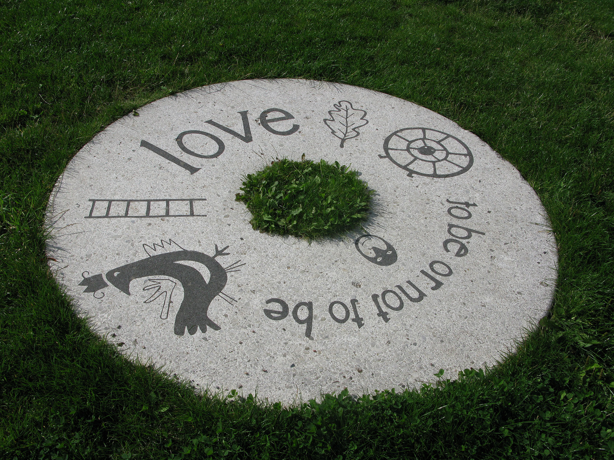 Love to be or not to be står det på en rund sten som är i gräs på marken. Flera symboler finns också på stenen som ett löv, en stege, en fågel och en rund ring