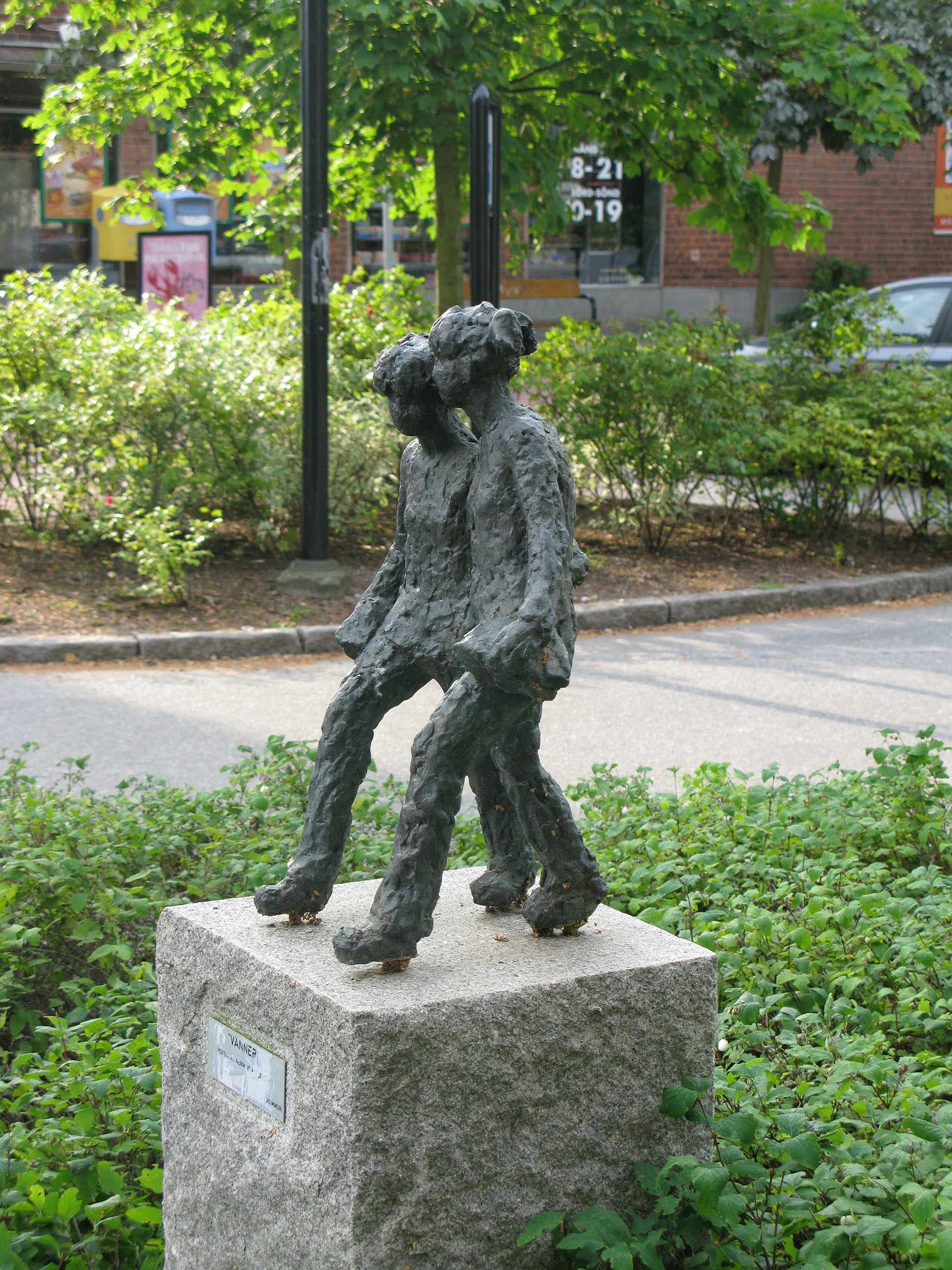 En skulptur i brons med två flickor som går tätt i hop. Grönska bakom.