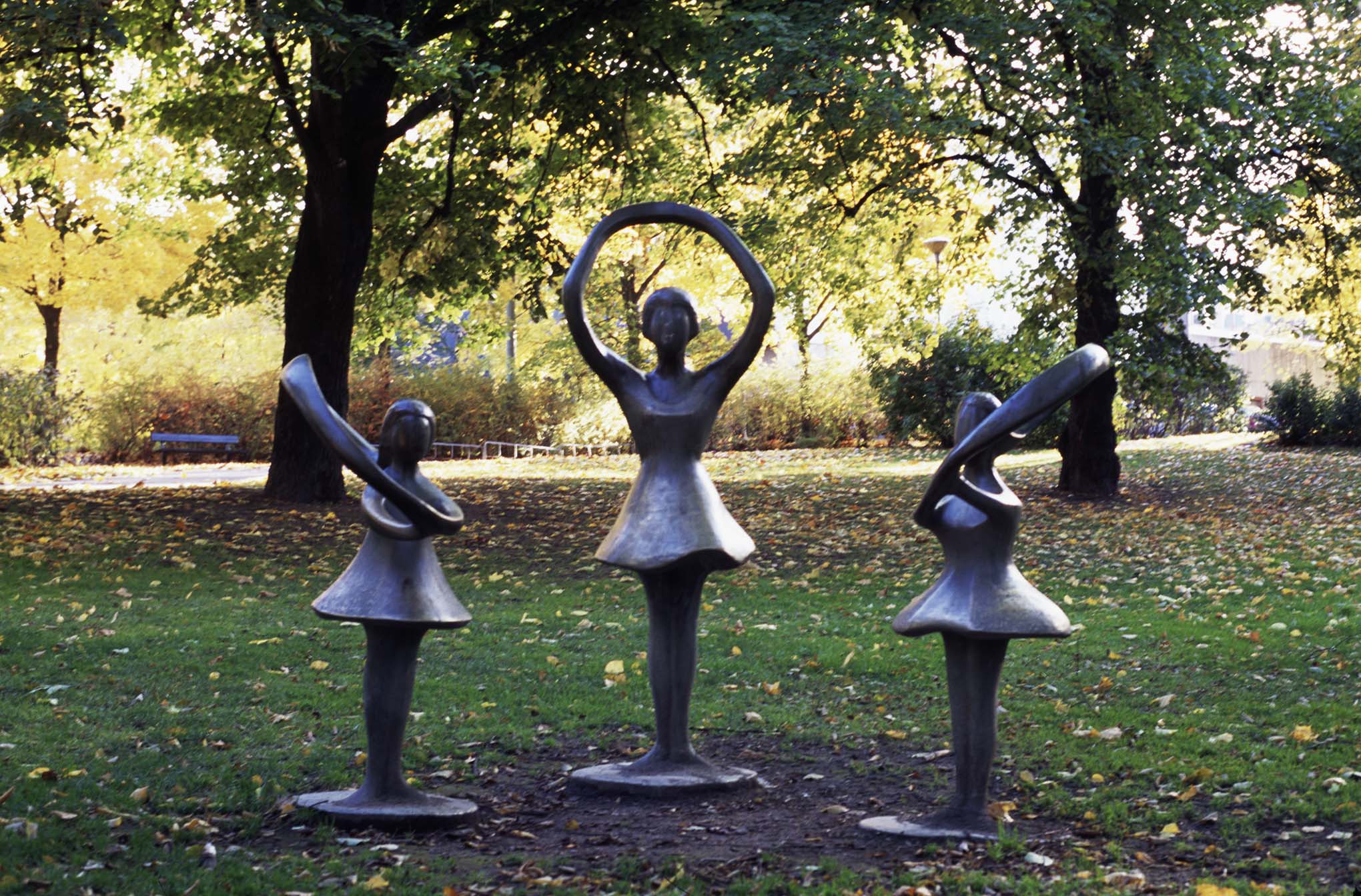 Tre skulpturer av flickor i brons med klockade kjolar står i en parkmiljö. Armarna är gjorda som runda ringar.