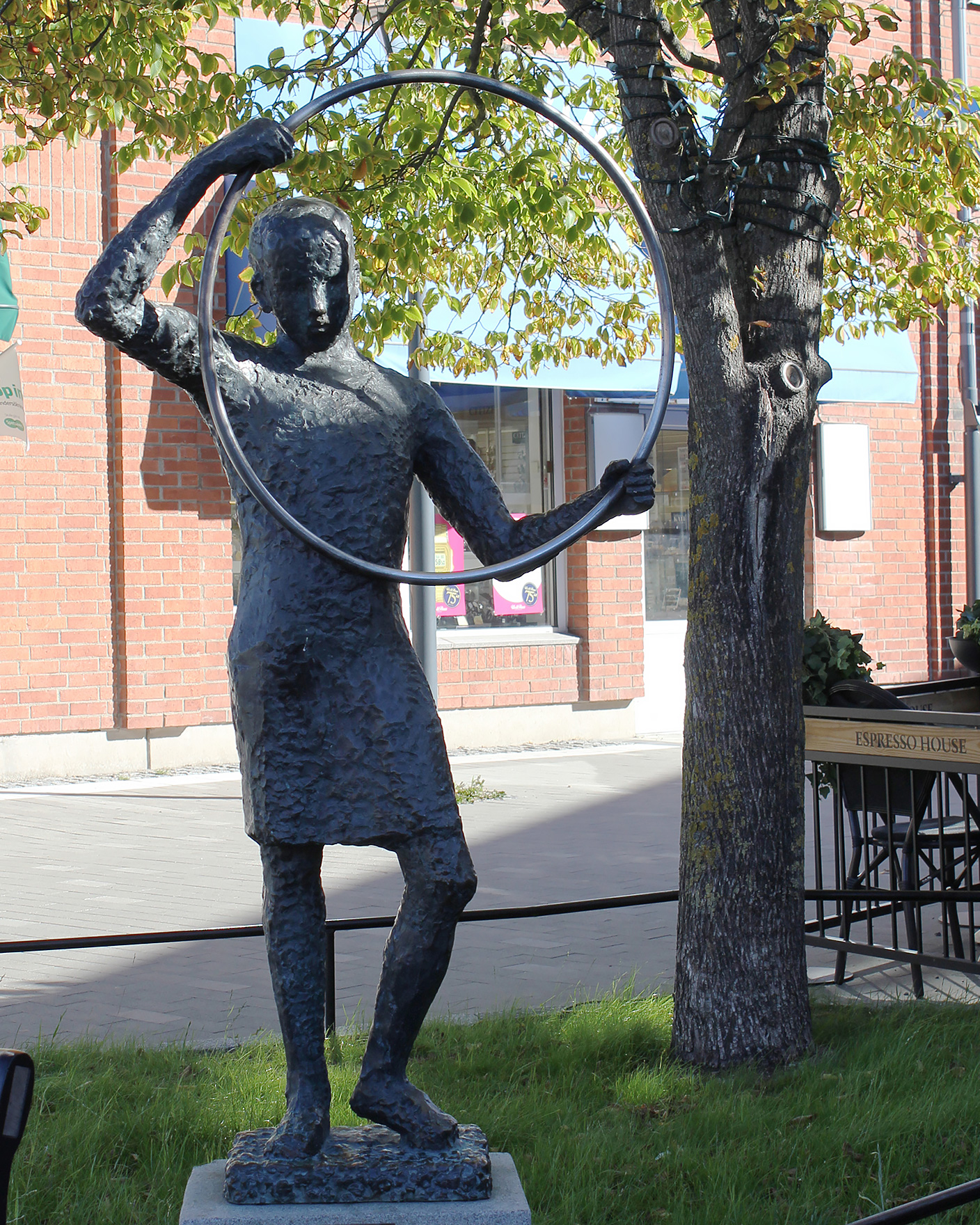 En skulptur av flicka som håller en metallring i sina händer. Träd, gräs och tegelhus bakom