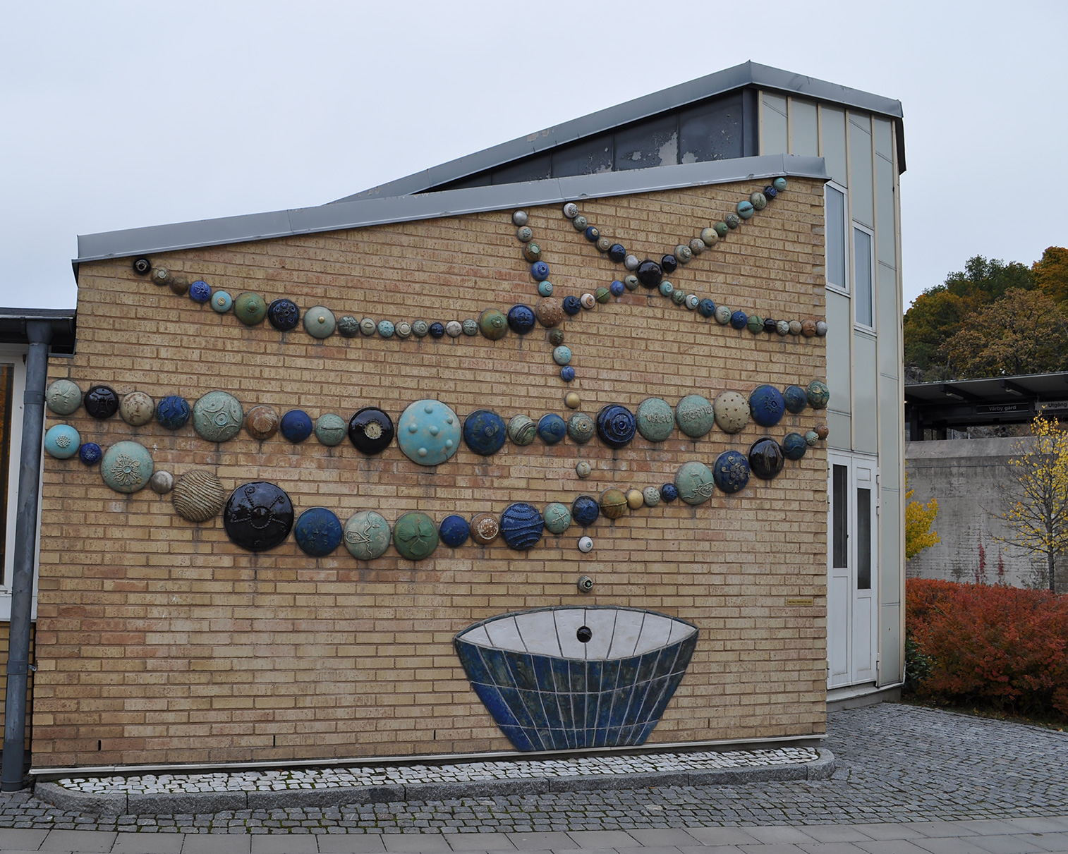 Runda klotformade figurer sitter på en vägg och bildar formen av ett halsband. Under står en blå skål.