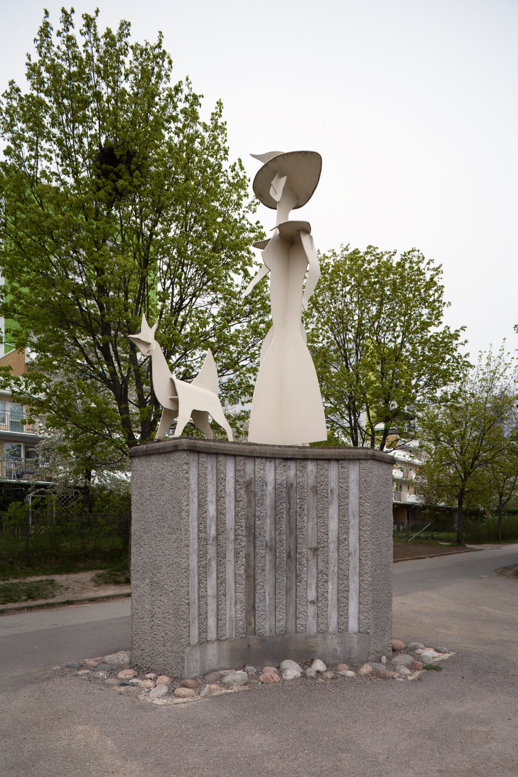 En vit skulptur av dam står på en hög grå sockel, framför sig har hon en vit hund.