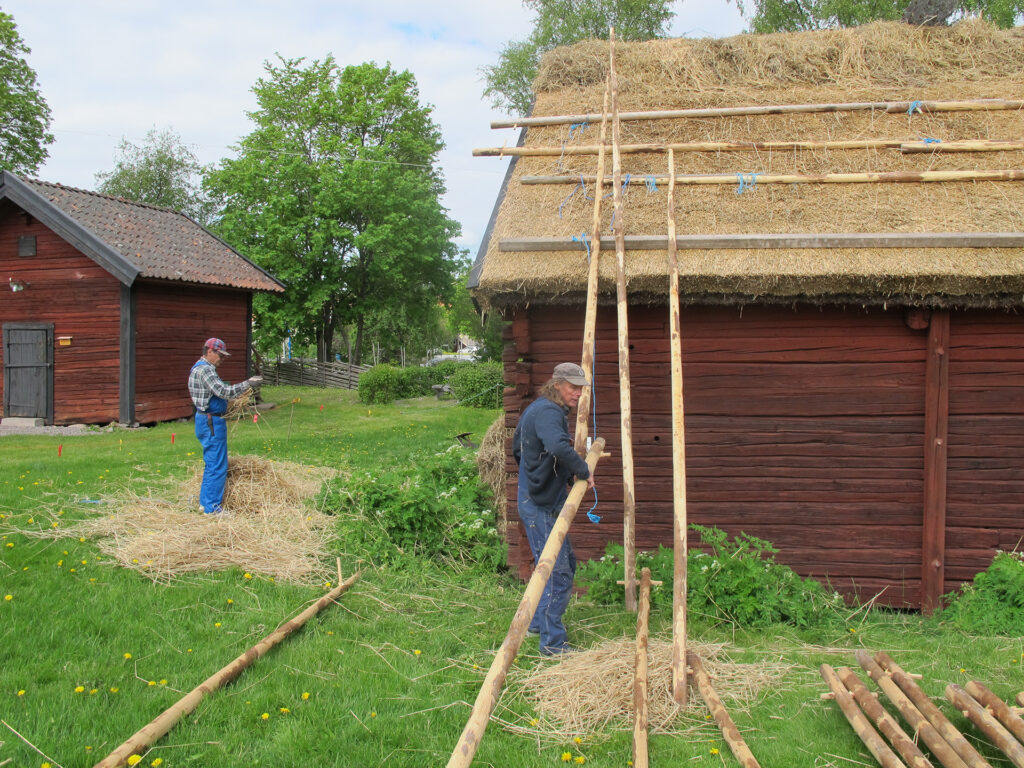 Två män arbetar med långa trästolpar som ska läggas på hus.