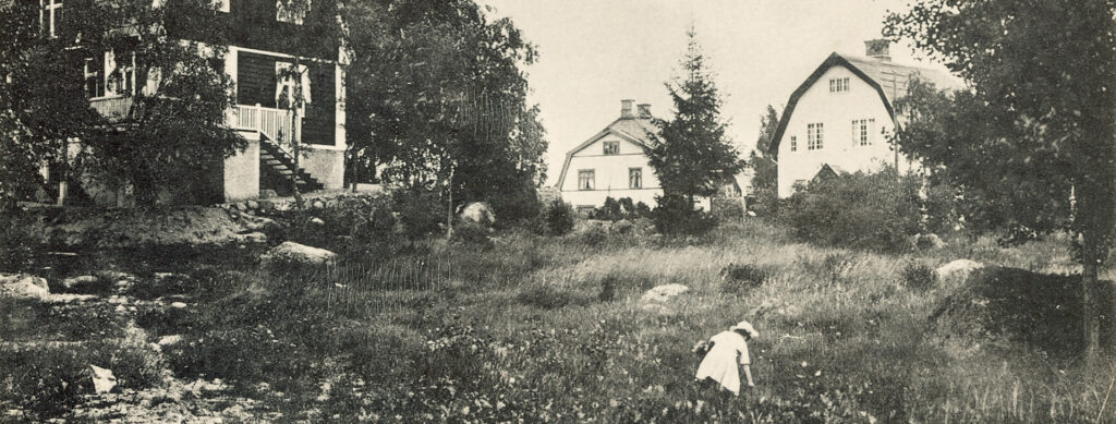 En flicka går och plockar blommor framför ett stort hus.