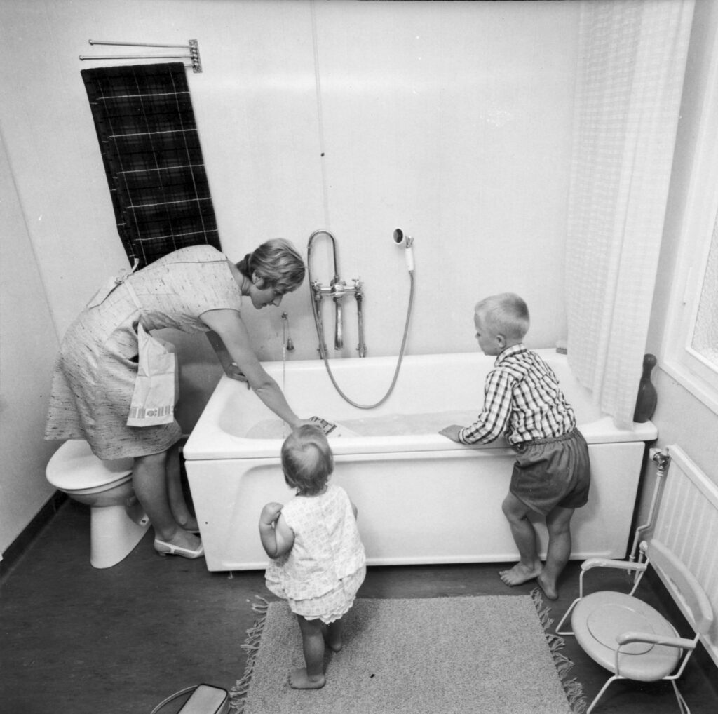 En kvinna står böjd över ett badkar fyllt med vatten. Två barn står bredvid.