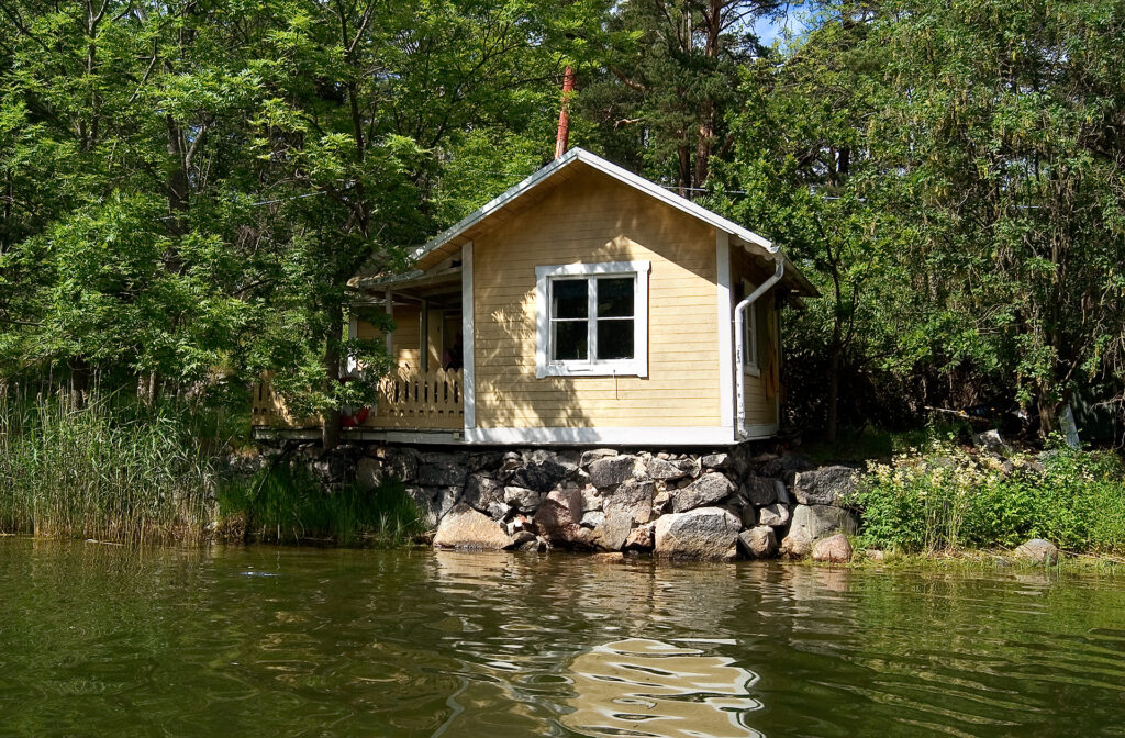 Ett litet hus som står i skogen, precis intill vattnet.