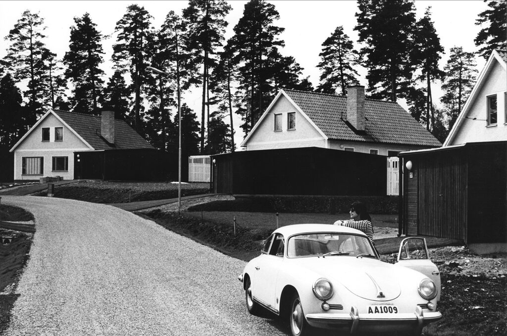 En gata med ett par hus bredvid. En bil står på gatan och en kvinna står och lutar sig mot bilen,