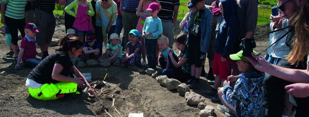 Museipedagog visar en skelettgrav för en grupp barn och vuxna vid Broby bro. 