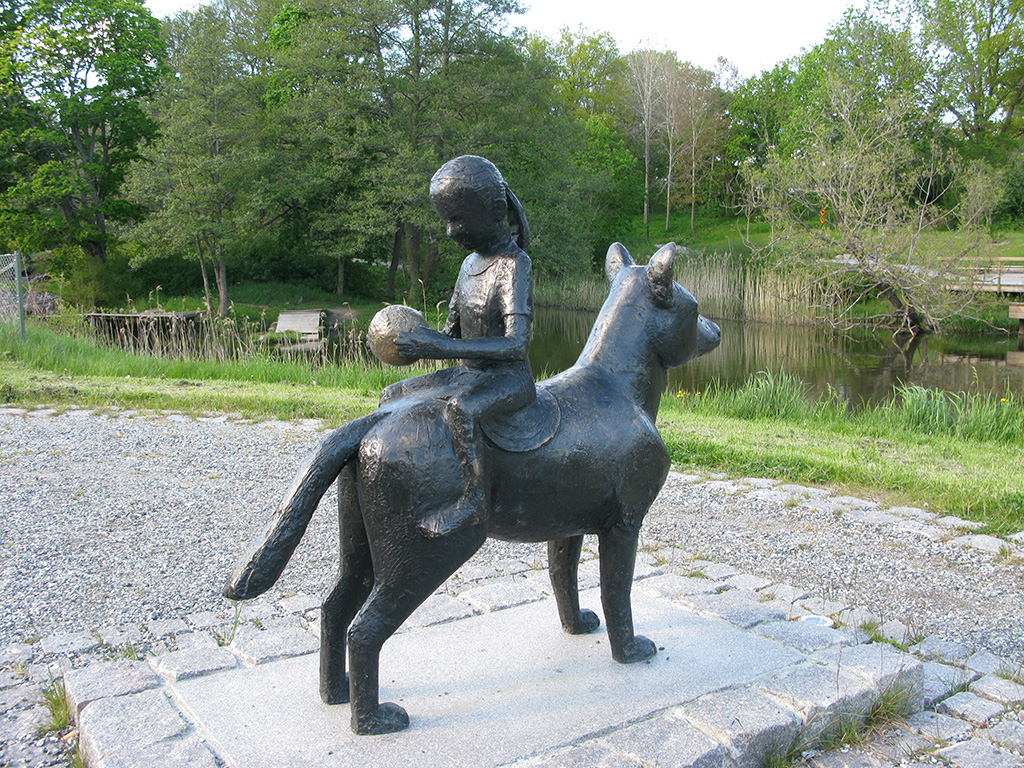 Skulptur i brons av en flicka som sitter på ryggen på en hund, vänd med ansiktet mot svansen.
