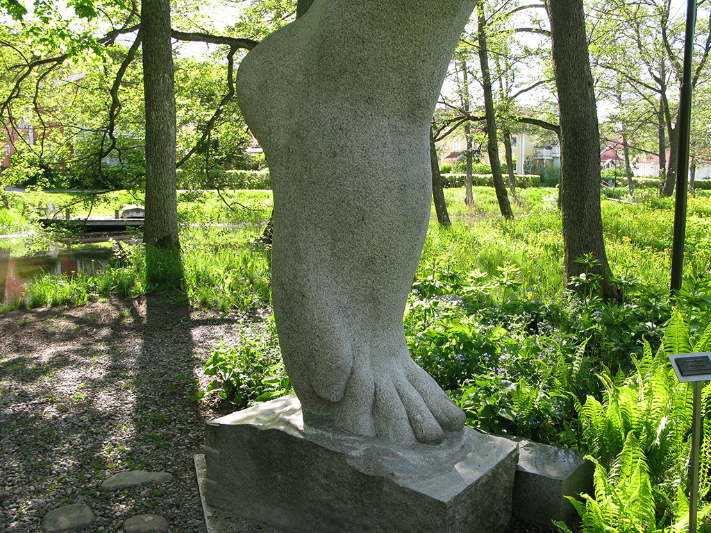 En skulptur av en jättestor fot i sten står på tå på en sten i skogen. Grönskande ormbunkar och träd runtomkring.