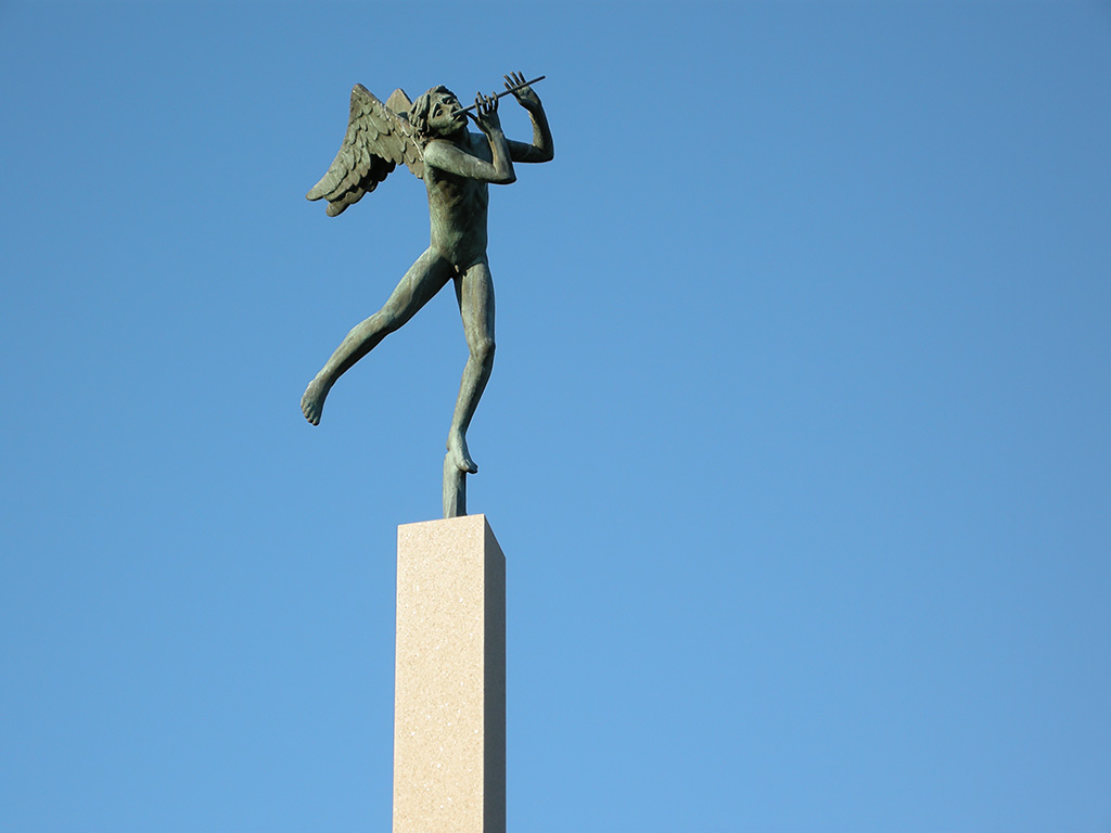 En skulptur av en pojke med vingar på ryggen som står på ett ben och heller håller en flöjt vid munnen. Skulpturen är på en hög sockel. Himmel bakom.