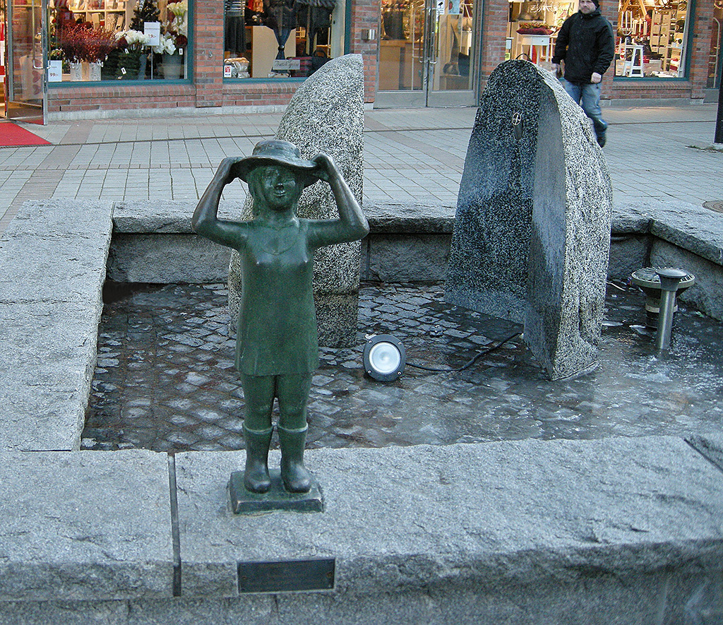 En skulptur av en som står och håller i en hatt på kanten till en fontän.