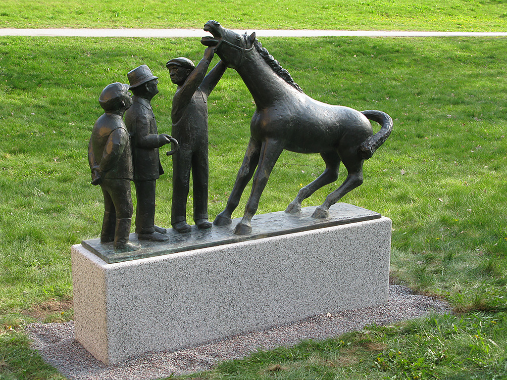 En skulptur med tre män och en häst på en ljusgrå sockel. En av männen håller i hästens grimma medan de andra tittar på. Hästen håller upp huvudet mot luften och har öppen mun. Bakbenen är böjda.