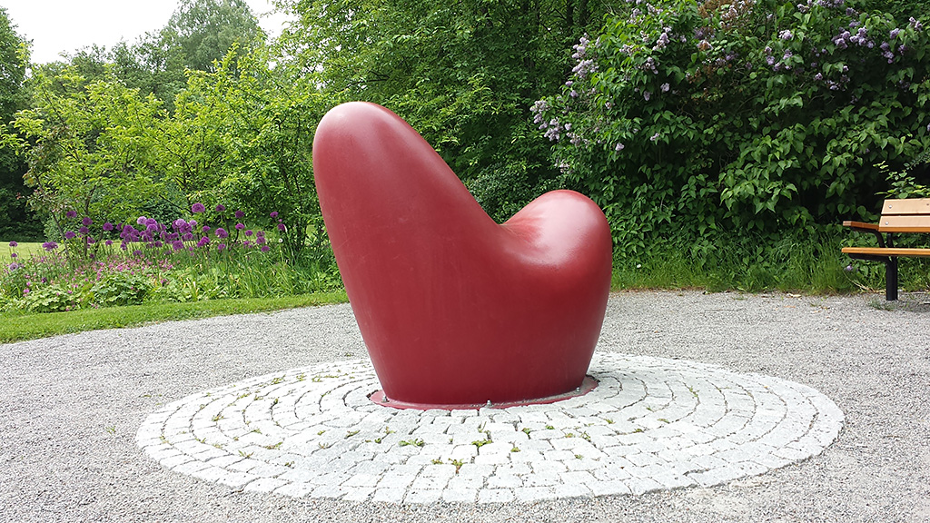 En röd abstrakt skulptur av en figur med rundade former. Står på en rundel av gatsten. Grönska bakom.