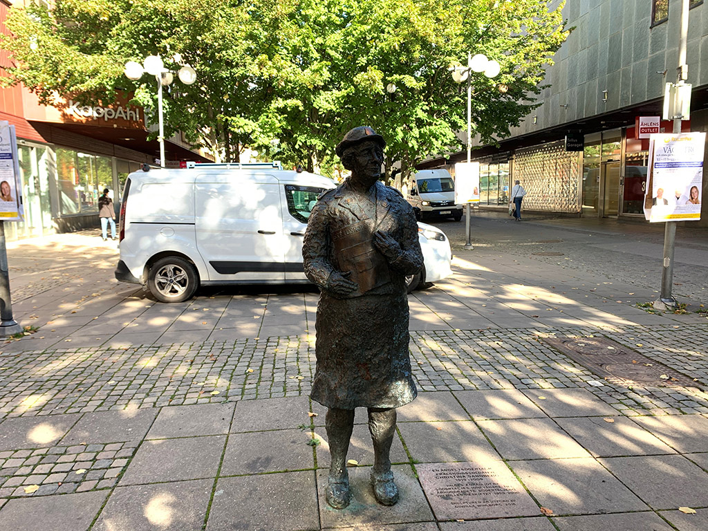 En skulptur av kvinna med hatt och dräkt i gatumiljö. Grönskande träd och en vit bil bakom.