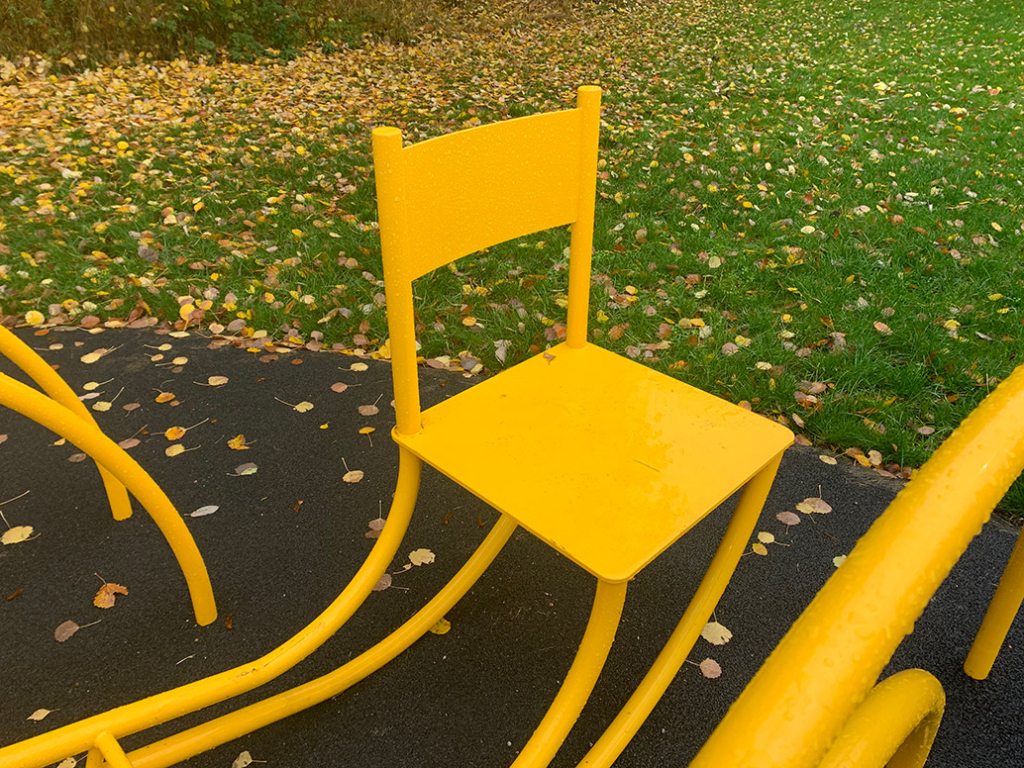 En gul stol men långa ben som står ute med gräs och gula löv bakom.