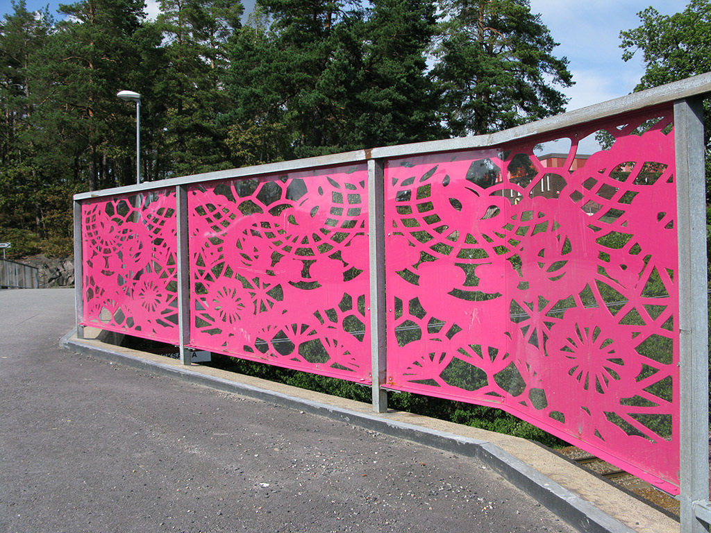 Ett bro med ett rosa broräcke med mönster som leder mot en skog.