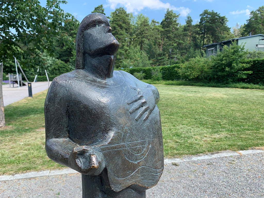 Bronsskulptur av gestalt som spelar gitarr och står på en grå sten.