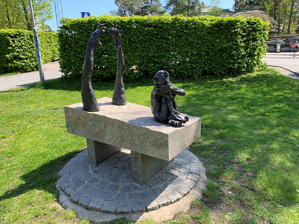 Skulptur i brons av en sittande person på en granitplatta med två ben som sticker upp bakom. Grönskande häck i bakgrunden.