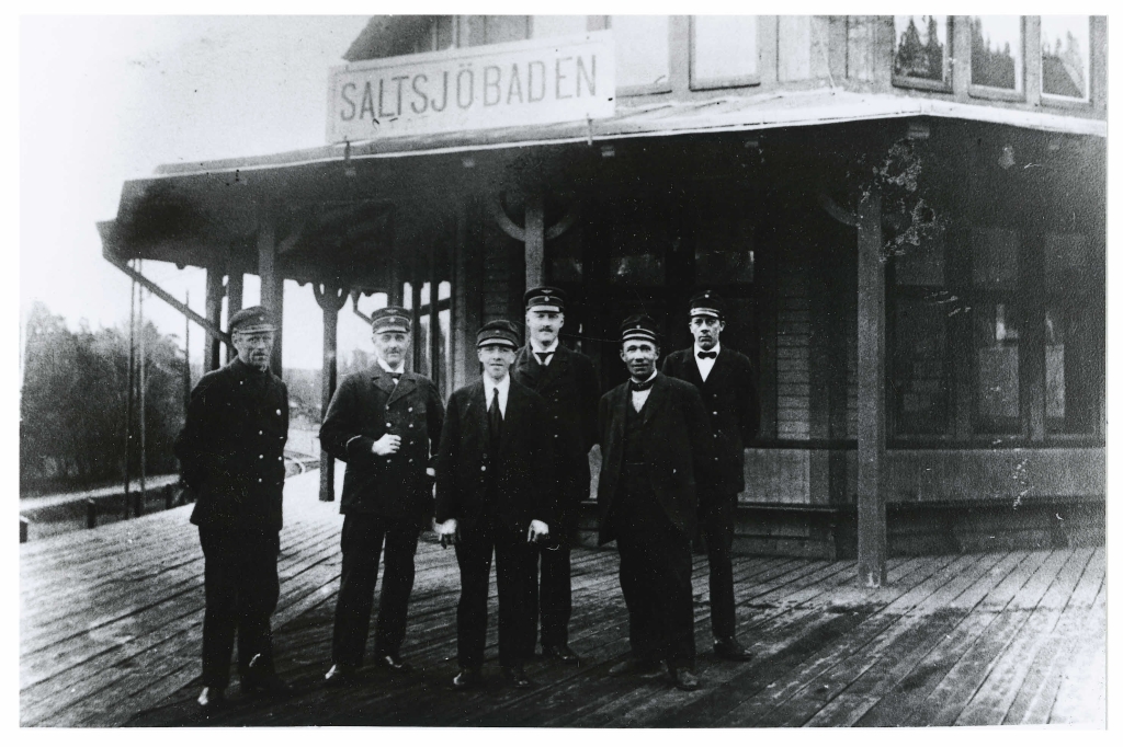 Svartvitt fotografi. 6 män i arbetskläder, uniform, står uppställda framför Saltsjöbadens gamla stationshus.