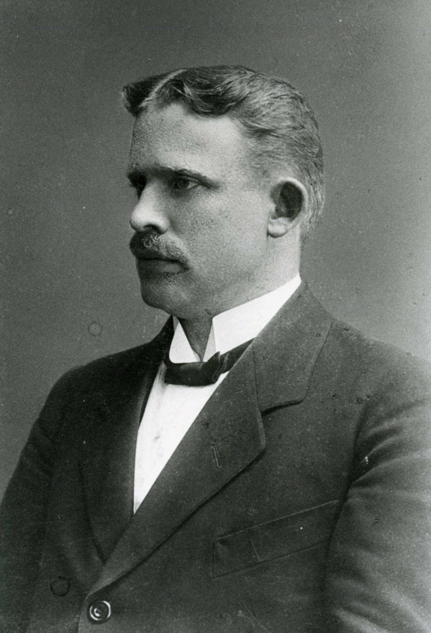 Svartvitt fotografi av en man i profil som bär fluga och mustasch.