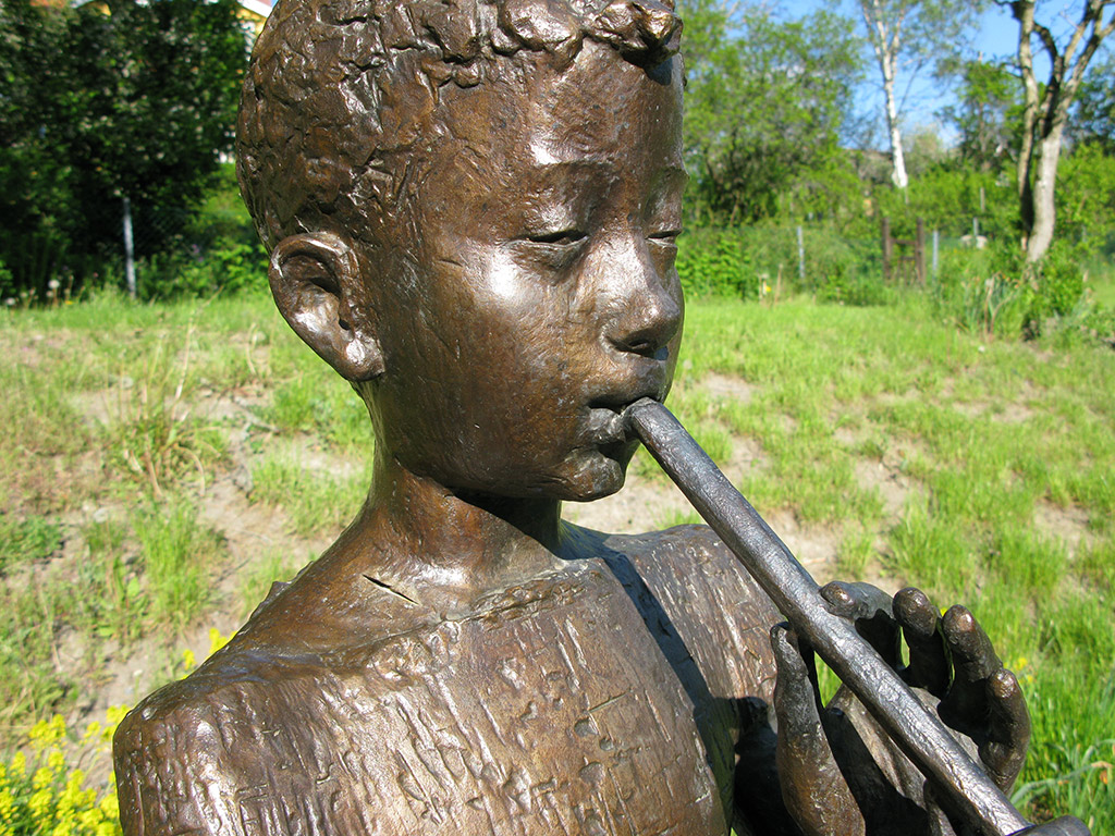 Skulptur av en pojke med flöjt i munnen. Grönska bakom.