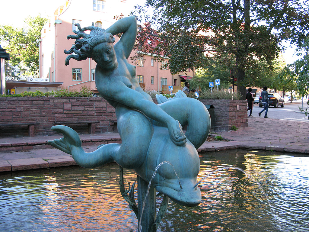 Skulptur av en kvinna på delfin i fontän. Hus och träd bakom