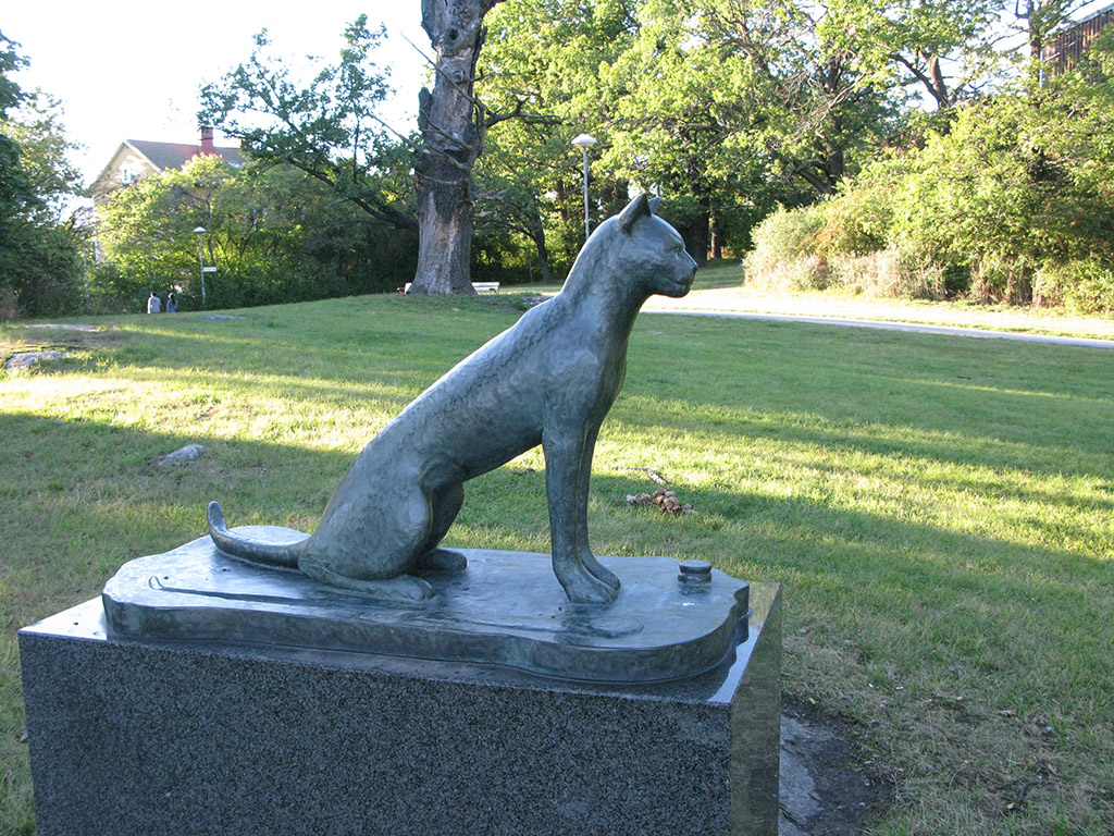 Skulptur i brons av sittande katt. Träd och gräs runtom.