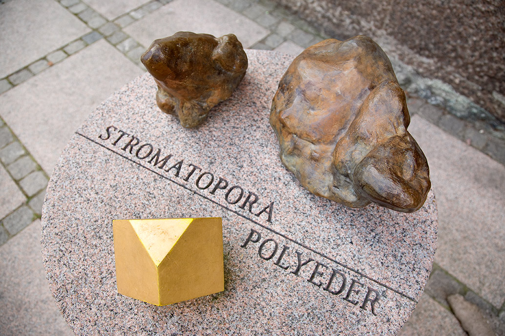 Tre abstrakta föremål på granit. Enkubisk form av mässing och två bubbliga bruna former. Inristad text i stenen som lyder Stromatopora Polyeder.