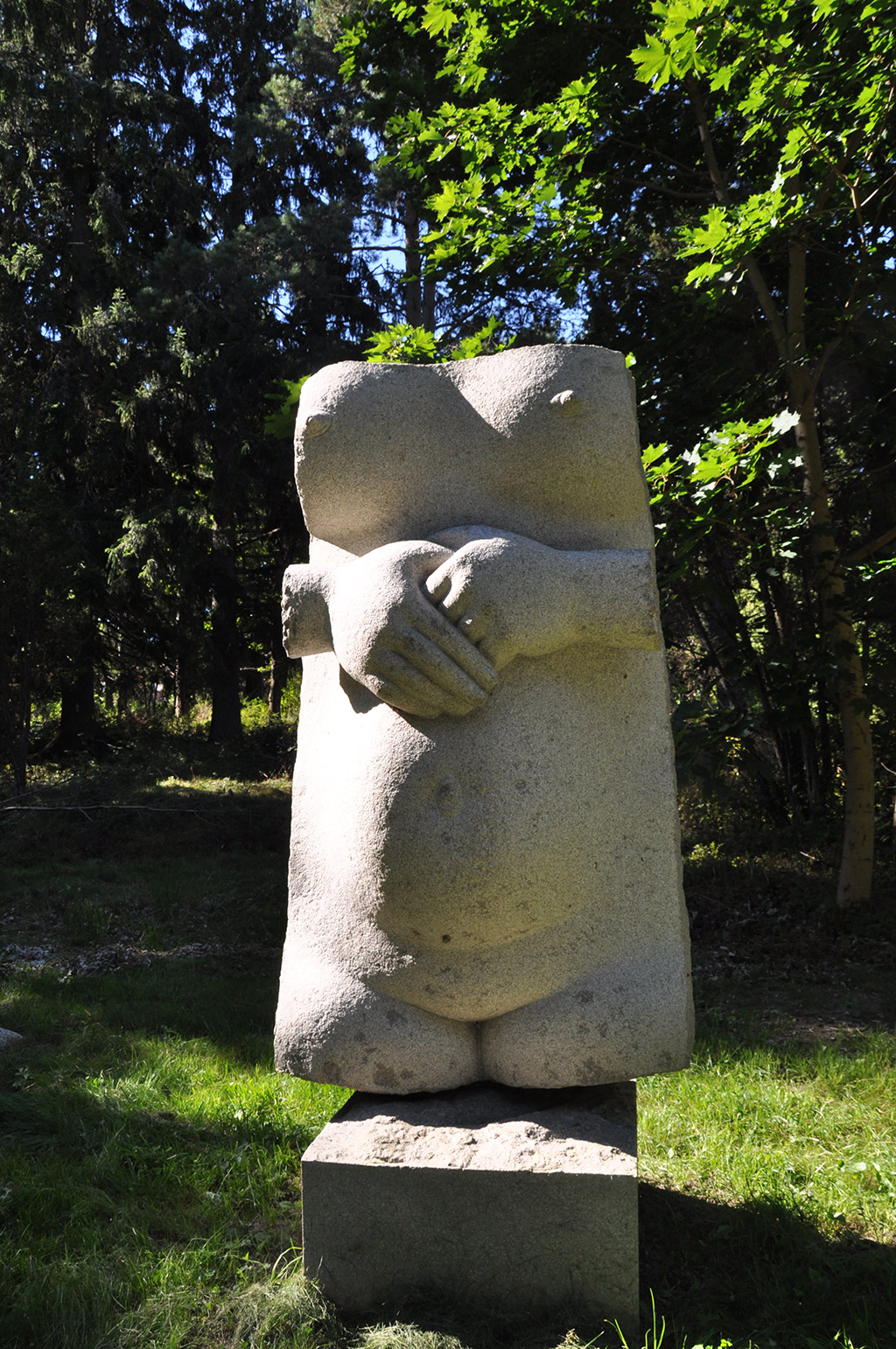 Skulptur av överkropp av kvinna i sten. Armarna korsade över magen. Grönskande träd och gräs.