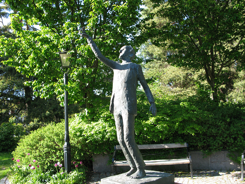 Bronsskulptur av stående man med ena handen lyft. Grönskande träd och bänk och gatlykta bakom.